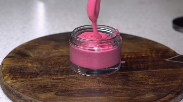 Woman placing pink homemade cream into a transparent glass jar on dark wooden background. Natural skincare, DIY beauty product, clean cosmetics, and wellness concept.