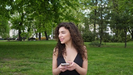 Fototapeta premium A beautiful stylish brunette woman sits on a lawn in the park in the summer and writes a message on her smartphone