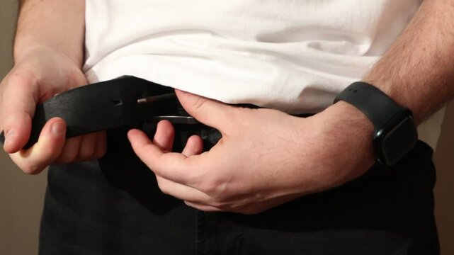 A man in a white T-shirt and black jeans fastens the belt on his pants