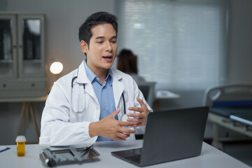 Asian doctor wearing lab coat and stethoscope is speaking during a video call with a patient using a laptop in a hospital room