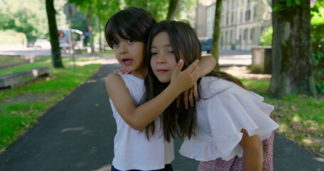 Brother and sister enjoying a warm day outdoors, hugging and smiling, with trees and a path in the background, both with long dark hair, creating a sweet family moment