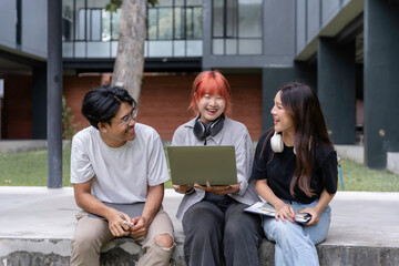 Three college students sitting outdoors, laughing and collaborating on a project using a laptop, showcasing teamwork and friendship