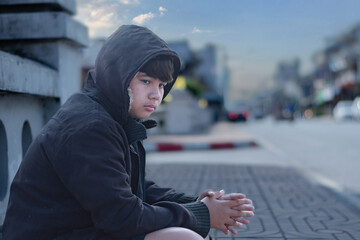 Asian young boy wearing a dark hooded jacket, sitting near a stone fence or barrier thoughtfully...