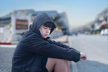 Asian young boy wearing a dark hooded jacket, sitting near a stone fence or barrier thoughtfully...