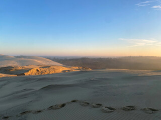 Sunrise over a desert of sand dunes with visible footprints trailing across the soft golden sand, creating a peaceful and serene atmosphere