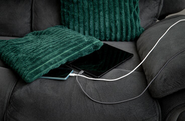 Smartphone and tablet computer are charging on the couch under a pillow in living room. Concept of overheat and fire hazard.