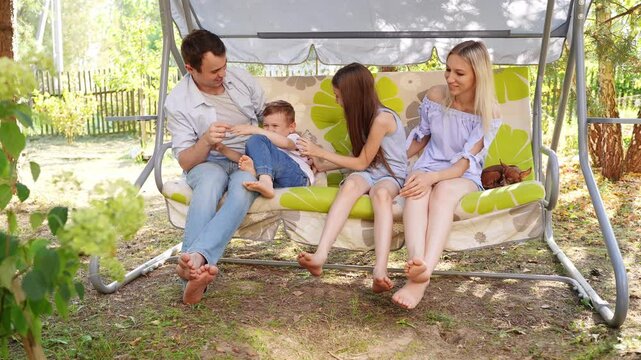 Cute family sitting together on swing in backyard, chatting, spending time together, enjoying rest in backyard. Mom, dad, brother, sister spend hot summer day in shade, tickling each other. Vacation.