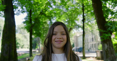 Naklejka premium Smiling girl with long hair standing on a tree-lined path in a sunny park, wearing a white blouse, enjoying the greenery and nature around, with buildings in the background