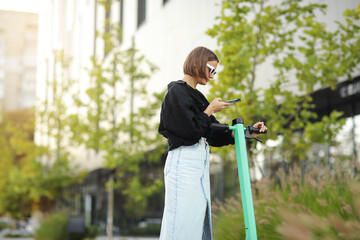 Stylish young woman with smartphone scans QR code for electric scooter rental. Girl using app on phone to unlock dockless transport on street. Urban style riding ecological vehicle. Modern technology