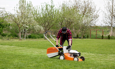 Man in casual clothes takes out bag with cut grass of lawn mower after mowing lawn with at own backyard. Husband takes care of garden. Modern gasoline garden equipment. Landscaping work at home