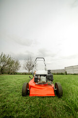 Gardening work tools. Wide angle view of orange electric lawn mower, wheels, motor on bright lush green grass at backyard. Rotary lawn mower machine cut lawn. Professional lawn care service.