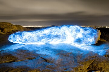 Nighttime landscape featuring a glowing blue rock.  A large rock, illuminated by bright blue light, sits atop a beige desert landscape.  City lights visible in the distance