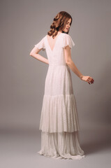 Fototapeta premium White shiny maxi gown with tiered skirt. Romantic portrait of smiling brunette woman in bohemian evening dress with short sleeves. Bridesmaid's fashion. Summer feminine look for hen party, rear view