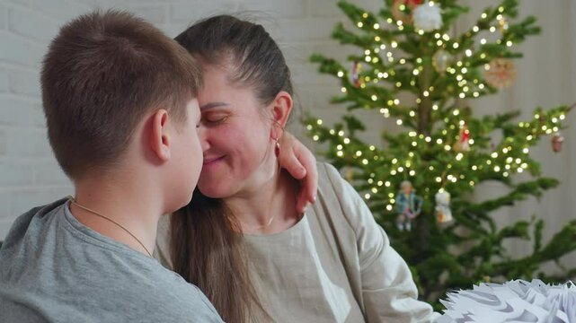 mother and son join heads together smiling warmly rubbing noses near christmas tree decorated with lights in cozy room expressing affection and emotional bonding during joyful festive season indoors