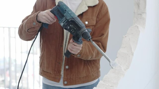 Unrecognizable construction worker demolishing concrete wall with rotary hammer, wearing protective safety gear. Renovation concept, closeup vertical view
