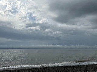 Obraz premium Cloudy sky over calm sea with small waves on a pebble beach. Several birds are flying in the sky. The weather appears overcast and humid.