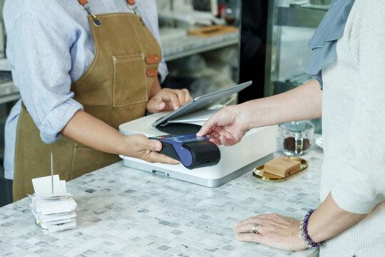 Asian senior female barista adult holding payment terminal while customer taps credit card at cafe demonstrating efficient cashless service in local small family coffee shop. Concept small business