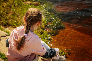 A young girl in a pink jacket sits quietly by the edge of a sunlit river, surrounded by fresh...