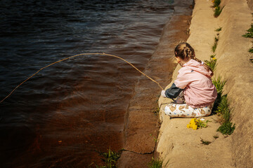A young girl in a pink jacket and patterned leggings sits by a riverbank, playfully fishing with a stick while holding dandelions nearby.