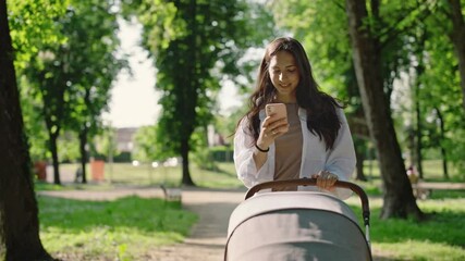 A beautiful young mother walks pushing a stroller with her baby in a park. The mother sends a message with her cell phone and smiles. Concept of Connection