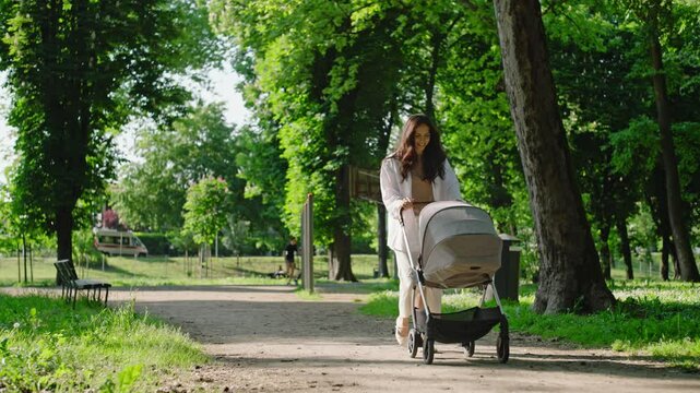 A beautiful young mother walks pushing the stroller with her baby in a park. On a sunset day the mother is happy to walk with her baby. Concept of family, freedom
