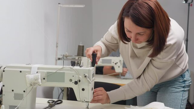 Professional garment designer loosens presser foot of electric sewing machine with screwdriver while another designer in background works on fabric using identical machine in shared workshop setting