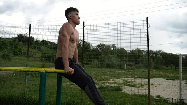 Strong man trains abdominal muscles raising legs on parallel bars at sports ground. Young shirtless sportsman exercising at nature. Sporty athlete working out at nature. Concept of sportive lifestyle