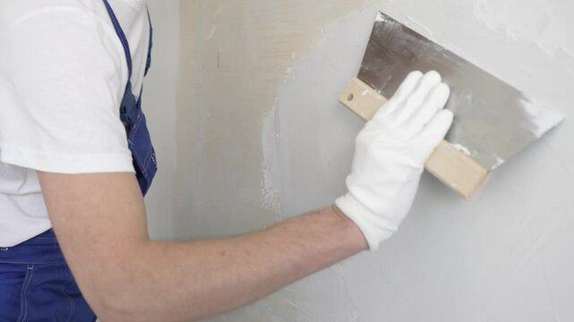 Unknown construction worker man plastering white wall, wearing blue overalls and protective white gloves during apartment renovation project, close-up