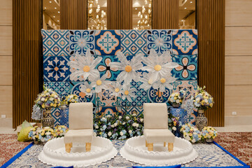 Elegant event setup with ornate tile backdrop, floral arrangements, and two chairs on platforms. Celebration, ceremony, or special occasion.