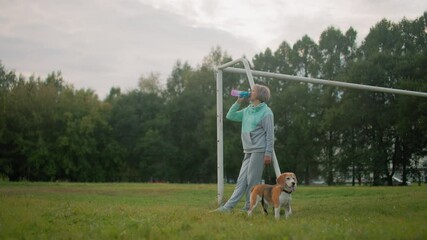 Sport woman dressed in gray and mint tracksuit resting on goalpost while drinking water from colorful bottle accompanied by her athletic beagle dog on grassy field during outdoor exercise session - Powered by Adobe