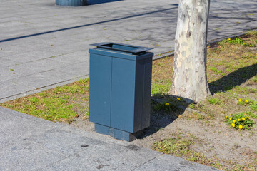 Gray Metal Waste Bin Placed Near Tree on Urban Sidewalk with Grass Patch in Spring