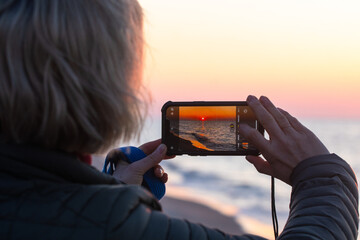 Close-up of a woman capturing a sunset with her smartphone—highlighting connection with nature,...