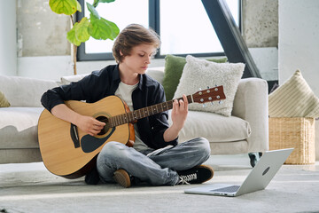 Teenage boy with acoustic guitar and laptop, having online music lesson