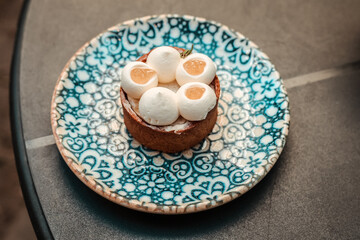 A top-down shot of a pastry tart served on a patterned blue plate, showcasing intricate design. The elegant dessert has white cream and fruit filling.