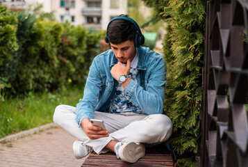 A young man with headphones using a mobile phone outside.