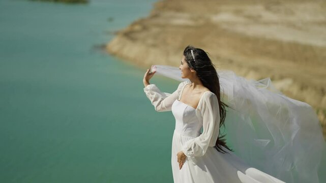 Woman in white dress near turquoise water on windy day, Elegant lady posing with flowing fabric by scenic cliffside lake view