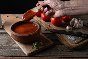 Hand holding wooden spoon with tomato sauce in setting with ingredients for preparation.