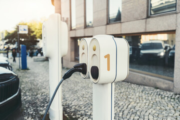 Close-up of an electric vehicle charging station in an urban setting, with a plugged-in charging cable, modern building reflections, and cobblestone sidewalk in soft evening light