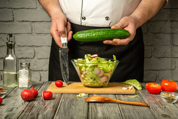 A chef is chopping cucumbers while preparing a fresh salad in a rustic kitchen. Various vegetables and ingredients are spread on the wooden table, creating a vibrant atmosphere