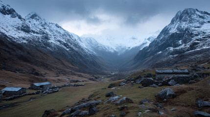 Dramatic mountain valley vista with snow capped peaks under a cloudy sky and rugged stone buildings in the foreground create a serene and remote landscape.