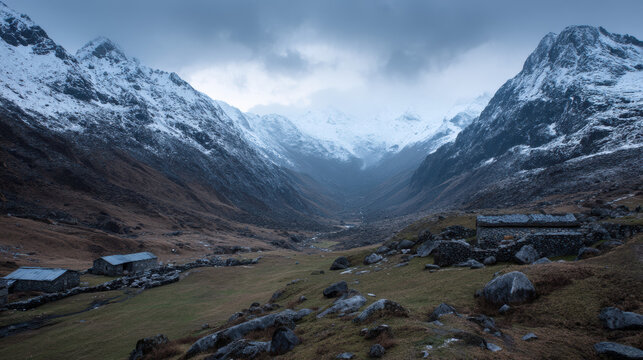 Dramatic mountain valley vista with snow capped peaks under a cloudy sky and rugged stone buildings in the foreground create a serene and remote landscape.