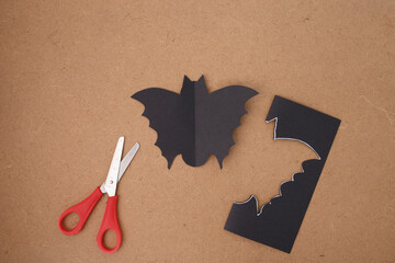 Cut bat silhouette with pointed ears and curved wings on brown background with red scissors, early...