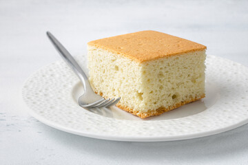 Slice of sponge cake on white ceramic plate with fork, close-up, minimal dessert concept, soft texture