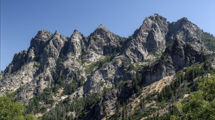 Jagged peaks of a rocky mountain range rise dramatically against a clear blue sky, showcasing nature's rugged beauty and scenic vistas on a sunny summer day.