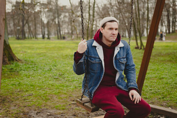 A young man sits calmly on a swing in a park surrounded by trees as autumn leaves cover the ground...