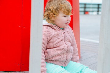 Fototapeta premium A young child dressed in a cozy sweater and light pants sits on a wooden structure at an outdoor playground The wind gently tousles her hair as she explores her surroundings