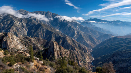 A breathtaking view reveals a magnificent mountain range under a clear blue sky, with clouds clinging to the peaks and valleys bathed in soft sunlight creating depth.