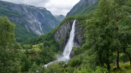 Fototapeta premium A beautiful waterfall cascades down a rocky cliff face surrounded by lush green trees and towe mountains under an overcast sky in the Norwegian wilderness.
