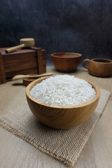 A wooden bowl filled with raw white rice placed on a piece of burlap over a wooden surface.