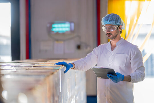 Food industry worker wearing hygiene gear inspects packaged products in a factory warehouse with warm sunlight in the background.